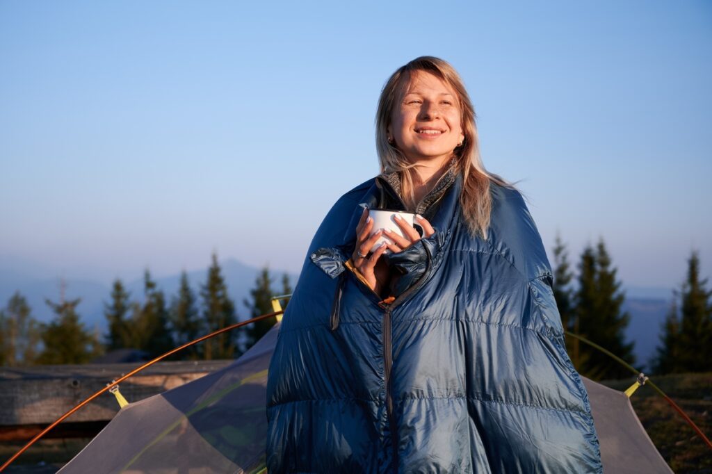 Cheerful woman hiker wrapped in sleeping bag holding cup of coffee. Cheerful woman hiker wrapped in sleeping bag holding cup of coffee.